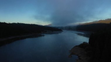 Sabahın erken saatlerinde sisli şafak ışıklarıyla bir gölün üzerinde uçmak. Hava aracı görünümü