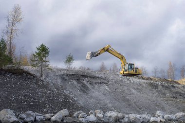 Sarı bir kazıcı bir tepede çalışıyor. Madencilik, yol çalışmaları, inşaat. Rotasyonel çalışma metodu, uzmanlık alanı. Düşük bulutlu gökyüzü.