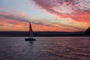Nehrin üzerinde güzel dramatik bir gün batımı. Karanlık sularda yelkenli. Uzakta, kıyıda. Mavi gökyüzü, kırmızı ve mor bulutlar.