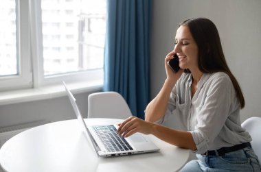 Girişimci kadın akıllı telefondan konuşuyor. Masada oturuyor ve dizüstü bilgisayar kullanıyor.