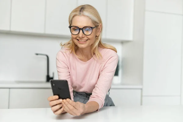 Щаслива усміхнена жінка середнього віку тримає смартфон, сидячи на кухні — стокове фото