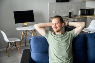 Gülümseyen bir adam kanepede dinleniyor, keyifle gülümsüyor, düşüncelerde kayboluyor.