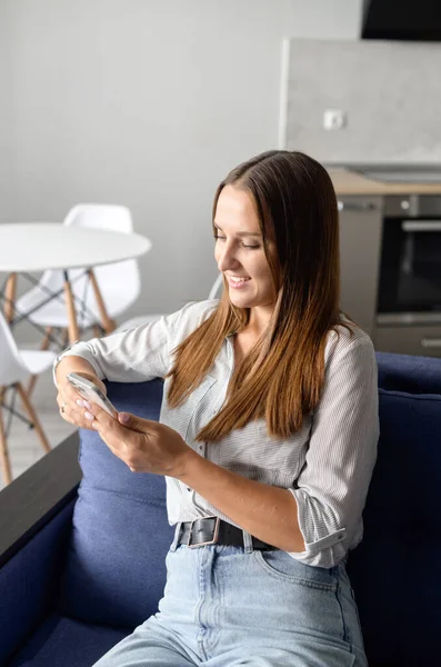 Mujer feliz en camisa casual usando teléfono inteligente sentado en el sofá en casa — Foto de Stock