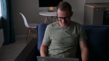 Dizüstü bilgisayar kullanan neşeli genç adam rahat koltukta oturuyor.