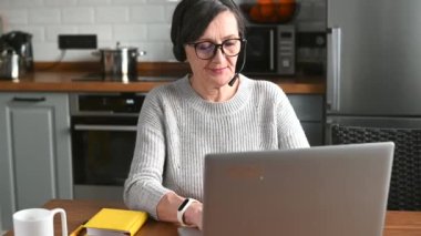 Kulaklık takmış yaşlı bir iş kadını, hesaplara bakarak konferans görüşmesiyle konuşuyor.