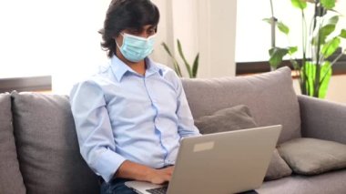Konsantre Hintli erkek serbest çalışan tıbbi maske takıyor kanepede oturuyor ve daktilo yazıyor.