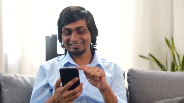 Serene sorrindo jovem indiano sentado no sofá em casa com um smartphone — Vídeo de Stock