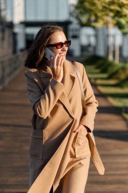 Sonbahar ceketli ve güneş gözlüklü bir kadın dışarıda yürüyor ve cep telefonuyla konuşuyor.