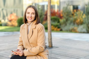 Neşeli, çekici genç bir kadın elinde moda akıllı telefonuyla dışarıda oturuyor.