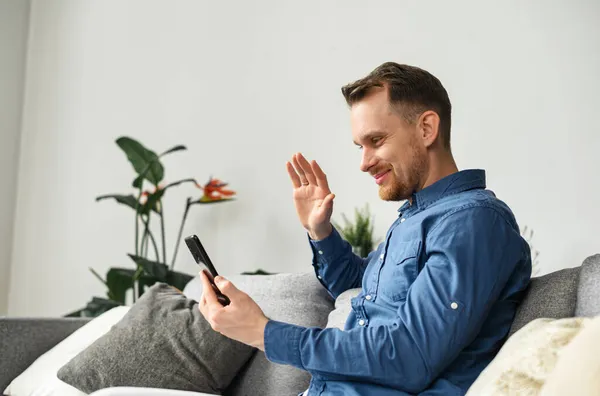 Позитивний молодий чоловік сидить на сірому дивані, дивлячись на телефон — стокове фото