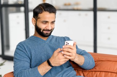 Odaklanmış sakallı Hintli adam evdeki kanepede uzanıyor ve elinde akıllı telefon tutuyor.