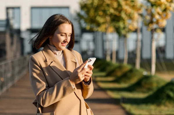 Mujer joven en camello abrigo de otoño de color caminando por la calle y utilizando el teléfono inteligente — Foto de Stock