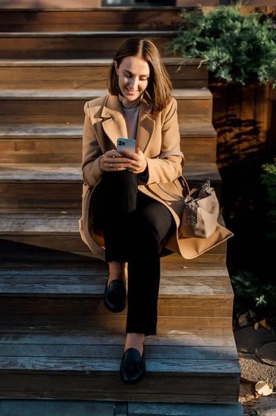 Retrato vertical de uma jovem sentada em degraus do lado de fora olhando para o celular — Fotografia de Stock