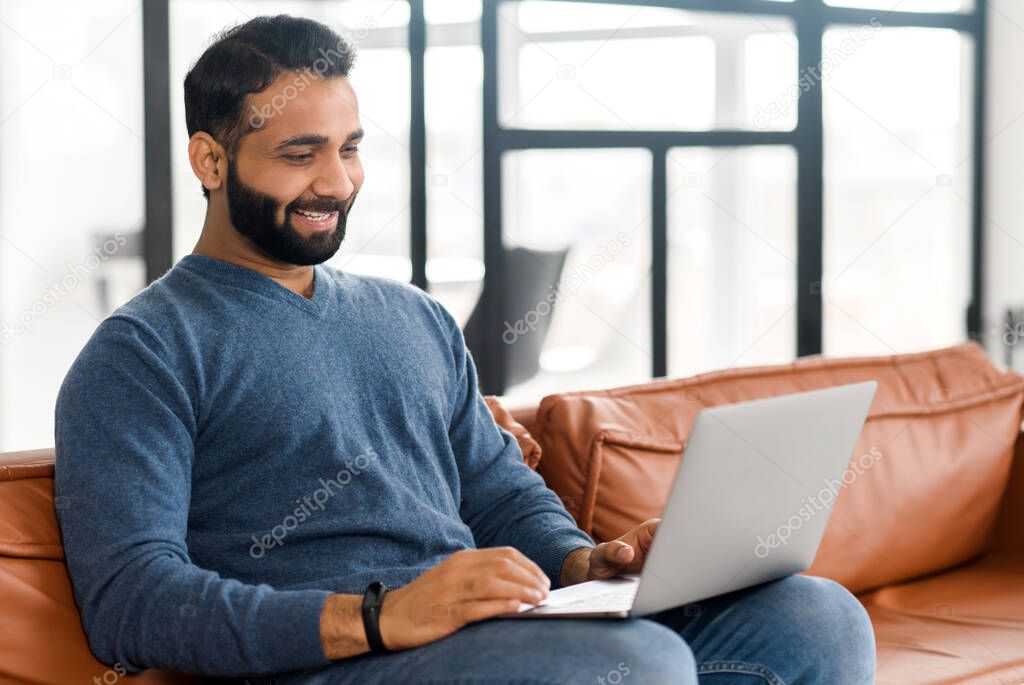 Cheerful Indian man wearing casual wear sitting on the sofa and using laptop