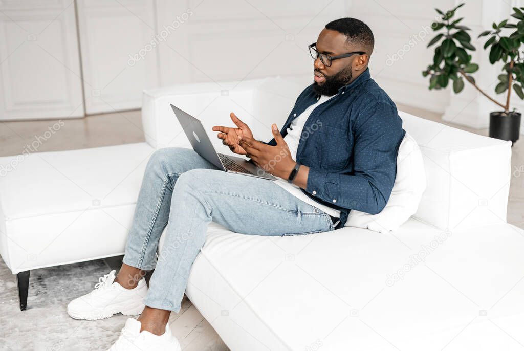 Focused African-American guy using laptop for video call sitting on the sofa at home