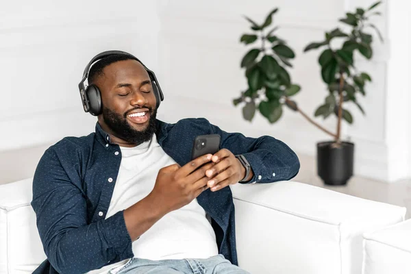 Chico africano guapo satisfecho usando teléfonos inteligentes y auriculares inalámbricos —  Fotos de Stock