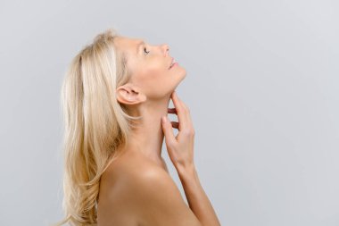 Çıplak omuzlu, çekici orta yaşlı bir kadın profilde duruyor, yukarı bakıyor ve gri arka planda boynuna hafifçe dokunuyor. Yaşlanma önleyici tedavi ve bakım. Spa ve rahatlama, kozmetoloji prosedürü
