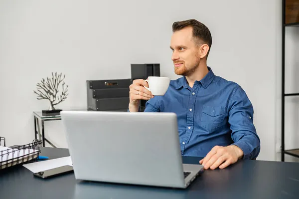 Χαμογελώντας αρσενικό υπάλληλο πίνοντας πρωινό καφέ, χρησιμοποιώντας ένα φορητό υπολογιστή — Φωτογραφία Αρχείου
