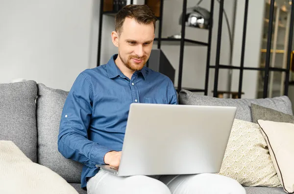 Усміхнений молодий бородатий чоловік використовує ноутбук вдома — стокове фото