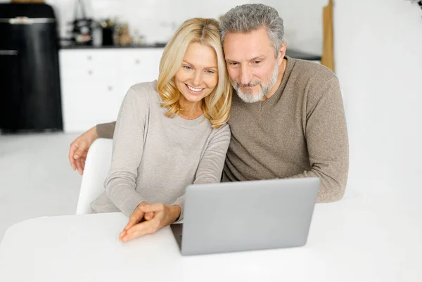 Σύγχρονη χαρούμενη ώριμος σύζυγος και η σύζυγος κάθονται στο γραφείο στην κουζίνα χρησιμοποιώντας φορητό υπολογιστή — Φωτογραφία Αρχείου