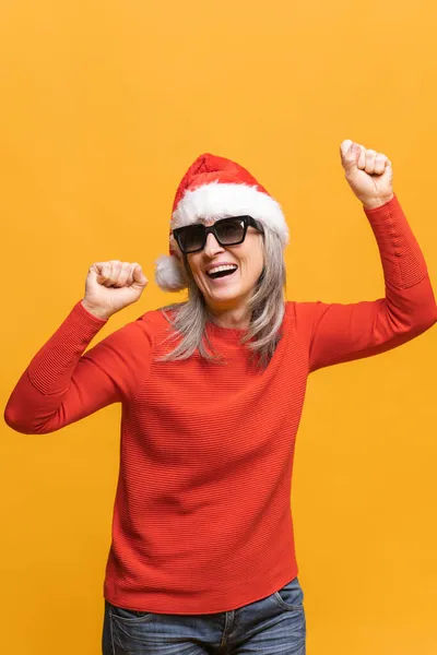 Щаслива середнього віку сіро-волохата жінка в червоному капелюсі Санта і сонцезахисні окуляри танцюють — стокове фото