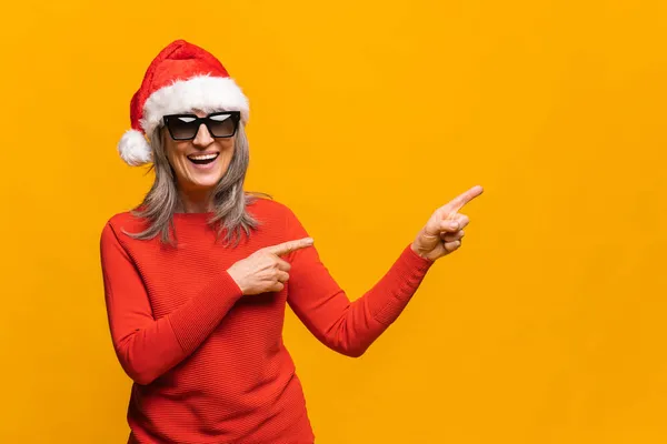 Усміхнена старша жінка в капелюсі Санта і сонцезахисних окулярах вказує пальцями вбік — стокове фото