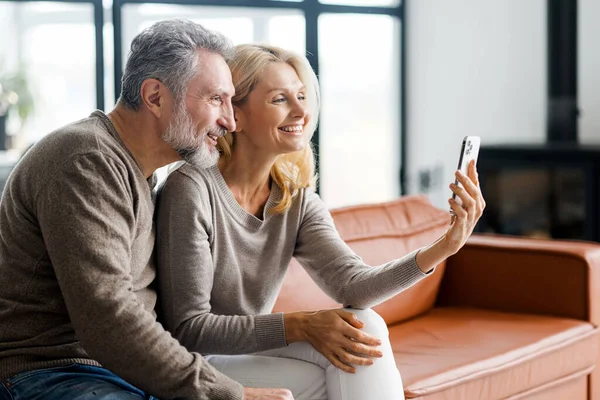 Lachend echtpaar van middelbare leeftijd met smartphone zittend op de comfortabele bank — Stockfoto