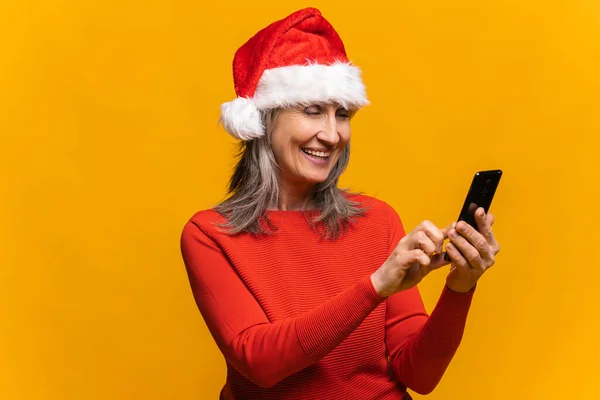 Wesoły atrakcyjny starszy kobieta w Santa kapelusz gospodarstwa smartfon — Zdjęcie stockowe