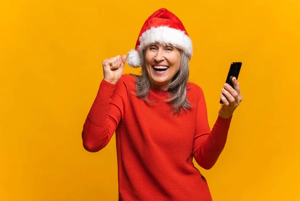 Mulher sênior feliz surpreso em Santa chapéu segurando smartphone e se alegra — Fotografia de Stock