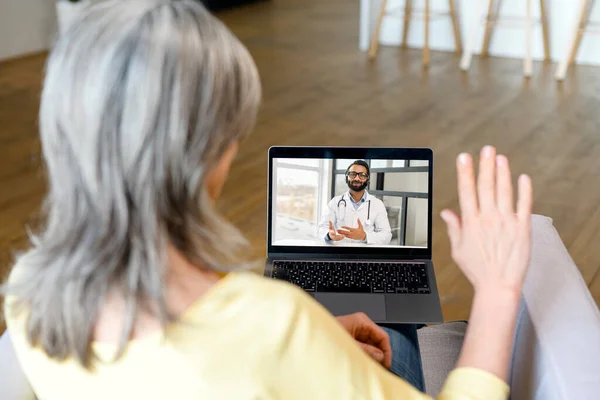 Зрелая пожилая женщина держит ноутбук и разговаривает с врачом онлайн из дома — стоковое фото