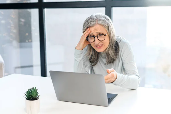Mulher sênior de cabelos grisalhos perturbada olhando para a tela do laptop sentado na mesa no escritório — Fotografia de Stock