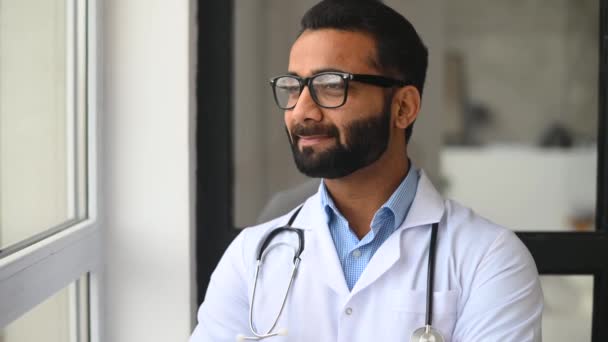 Indiase mannelijke arts kijkt door het raam bedachtzaam, dan draaien en kijkt naar de camera — Stockvideo