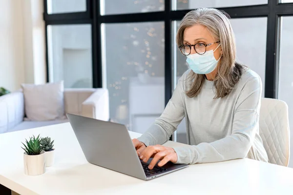 Сіро-волохата зріла бізнес-леді носить захисну медичну маску з ноутбуком — стокове фото