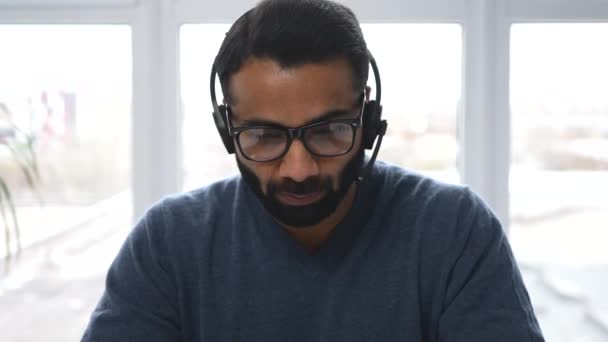 Front view of mid-age Indian male in casual wear using headset and modern laptop — Stock Video
