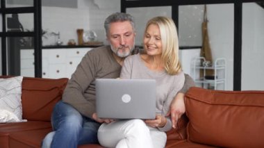 Orta yaşlı aşık çift evdeki kanepede dizüstü bilgisayar kullanıyor.