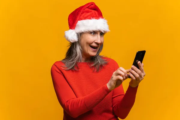 Zastanawiam szczęśliwy szczęśliwy Dojrzałe lady noszenie Santa kapelusz patrzeć na smartfona — Zdjęcie stockowe