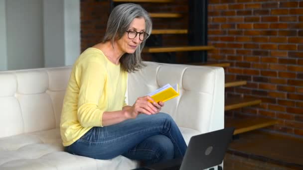 Mujer madura enfocada viendo webinars en línea en el portátil sentado en el sofá — Vídeos de Stock