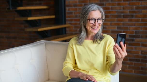 Positiv senior moden dame i en gul jumper taler online på telefonen, videoopkald – Stock-video