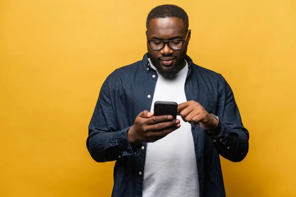 Foto van aantrekkelijke bebaarde zwarte man in casual shirt met behulp van smartphone geïsoleerd over grijze achtergrond, raakt pad scherm, noodzakelijk hulpmiddel voor de productiviteit, vindt inspiratie online — Stockfoto