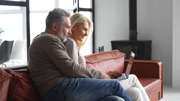 Pasangan setengah baya yang ceria dengan laptop duduk di sofa kulit yang nyaman — Stok Video