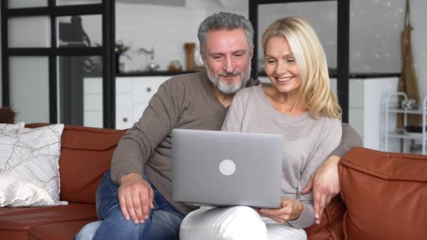 Despreocupados cónyuges de mediana edad utilizando el ordenador portátil sentado en el sofá en la sala de estar moderna — Vídeos de Stock
