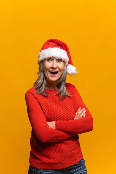 Radostná starší šedovlasá žena v Santa klobouku stojí se zkříženýma rukama a dívá se do kamery — Stock fotografie