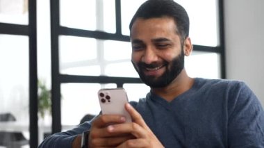 Kanepede oturan neşeli orta yaşlı Hintli adam akıllı telefon kullanıyor.