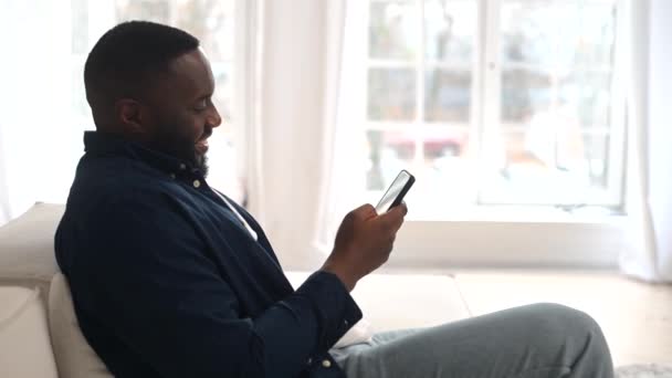 Atraente millennial barbudo afro-americano homem relaxante e sentado no sofá confortável — Vídeo de Stock