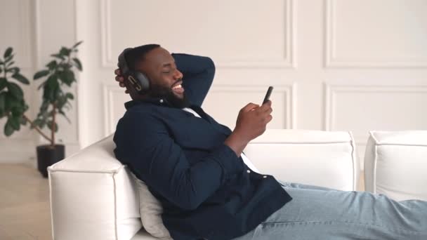 Satisfeito cara Africano está gostando de música deitado no sofá — Vídeo de Stock