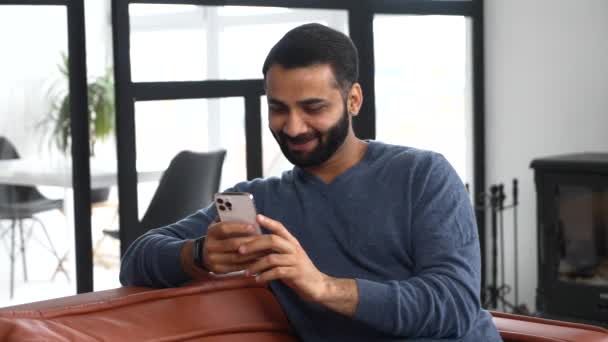 Спокійний усміхаючись індійському чоловікові, який сидить вдома на дивані, тримає смартфон. — стокове відео