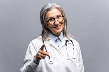 Gülümseyen, gri saçlı, beyaz elbiseli, gri elbiseli kadın doktor.