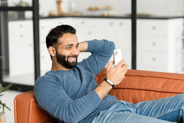 Hilarante hombre indio de 30 años usando un teléfono inteligente sentado en el sofá en casa — Foto de Stock