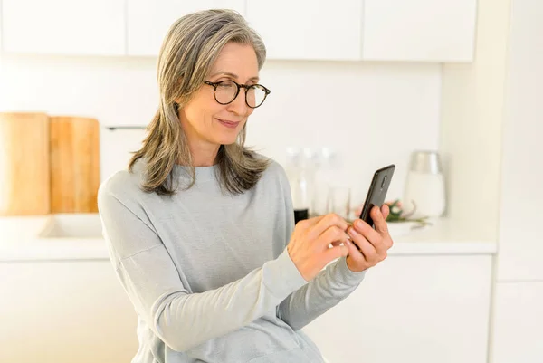 Šťastná usměvavá dospělá žena drží smartphone sedí v kuchyni — Stock fotografie