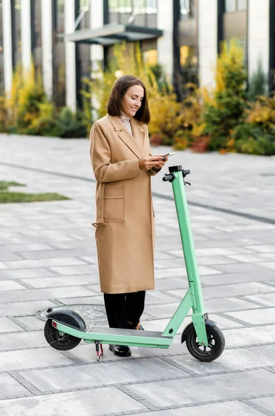 Donna felice in cappotto autunnale utilizzando smartphone per affittare scooter elettrico moderno — Foto Stock